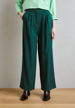 Pantaloni a gamba larga in tessuto verde scuro, caratterizzati da pince e tasche laterali, abbinati a stivali con tacco marroni. Sfondo neutro.