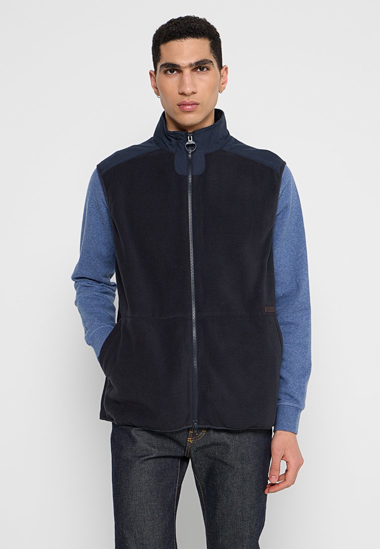 Homme portant un gilet en polaire noir zippé par-dessus une chemise à manches longues bleue et un jean foncé, se tenant devant un fond clair uni.
