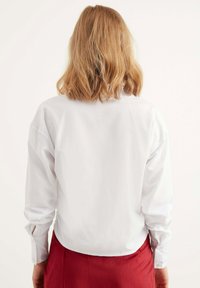 Camicia bianca con bottoni e maniche lunghe, caratterizzata da un retro leggermente accorciato e polsini con chiusure a bottone, indossata con una gonna plissettata rossa.