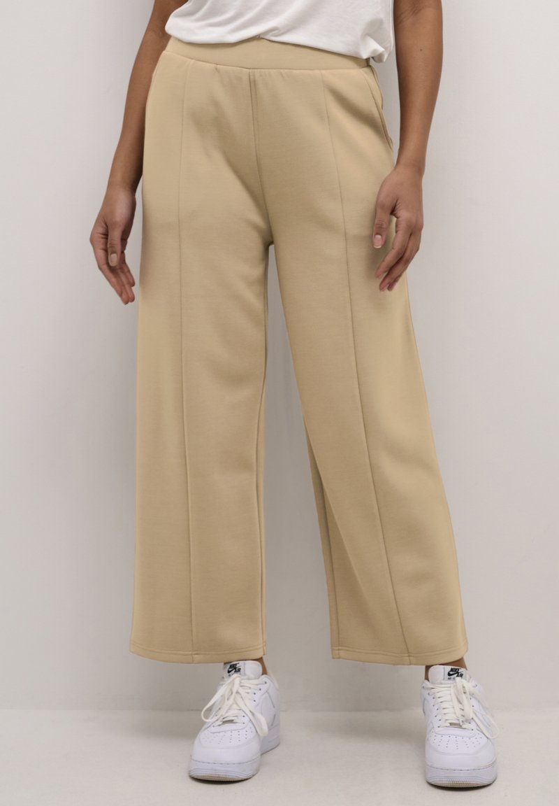 Oseba v bež širokih hlačah in belih Nike supergah stoji ob enostavni svetli steni.