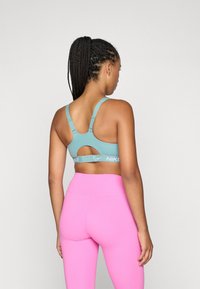 Svijetloplava sportska brassiere s izrezom, podesivim trakama i kontrastnim sivim Nike logotipom. U paru s visokim ružičastim tajicama.