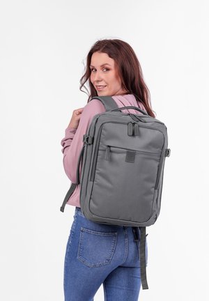 Šedý batoh s přední kapsou, nastavitelnými popruhy a polstrovaným zadním dílem, nošený na osobě. Materiál má hladkou texturu a minimální akcenty.