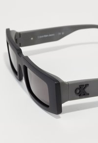 Ochelari de soare cu un cadru gri mat și lentile întunecate. Prezintă un logo negru embossat pe lateral. Design rectangular și voluminos.