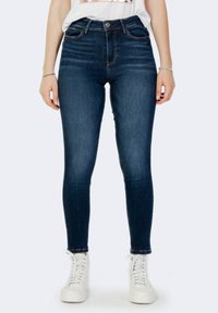 Jeans de mezclilla de tiro alto, ajustados, en azul oscuro con una textura suave, diseño de cinco bolsillos y detalles de costura. Combinados con zapatillas blancas.