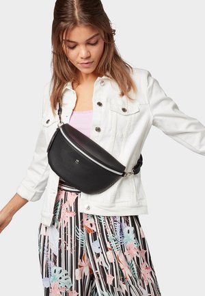 Junge Frau, die eine weiße Jacke, gestreifte Blumenhose und eine schwarze Umhängetasche mit silbernem Reißverschluss trägt und nach unten schaut.