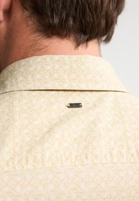 Camisa amarilla con estampado de diseño geométrico, con una pequeña etiqueta metálica en el cuello de la parte posterior. Textura de tela suave.