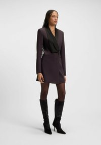 Abito corto in tessuto viola scuro, maniche lunghe, scollo nero a scallop, e silhouette aderente, abbinato a stivali neri con tacco al ginocchio.