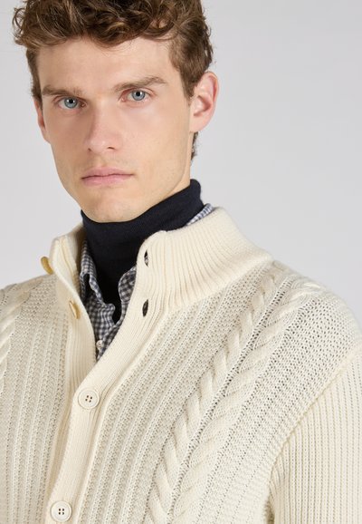 Crème kabelgebreide cardigan met een geribde kraag en knoopsluiting. Draagt over een marineblauw turtleneck en geruit shirt, met een getextureerd ontwerp.