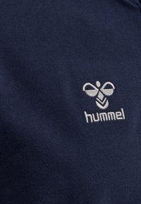 Marineblå tekstil med et broderet hvidt logo, der har et stiliseret bi-design, og brandnavnet "hummel" nedenunder.