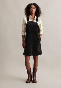Robe salopette en denim noir avec des bretelles réglables, une poche avant et une coupe évasée, associée à une chemise à carreaux légère et des bottines brunes.
