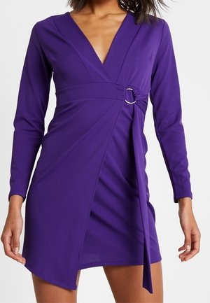 Femme portant une robe portefeuille violette à manches longues avec un détail de bague argentée à la taille et un ourlet asymétrique.