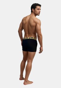 Svarta boxershorts med en gul midja som har texten "UKK", mjukt tyg och en tajt passform, visad från en bakre vinkel.