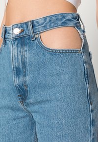 Jeans de denim azul de cintura alta com detalhes cortados nas laterais, costura laranja e um único fecho de botão prateado.