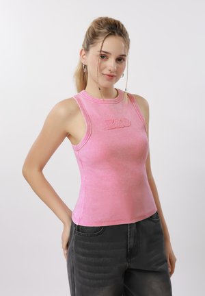 Jeune femme aux cheveux blonds portant un haut rose sans manches avec le logo "E&O", vêtue d'un jean foncé, posant devant un fond clair uni.