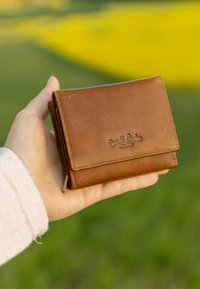 Rudas odinis piniginė su tekstūruota paviršiumi, turinti sagtį uždaryti ir iškilmiu tekstu "Gusti Leder studio" priekyje.