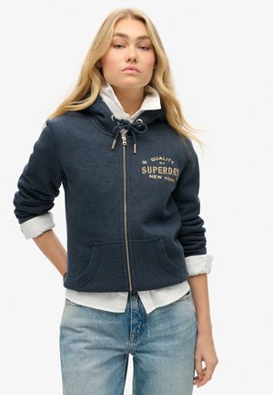 Navy zip-up hoodie with a drawstring hood, front pockets, and gold embroidered logo. Worn over a white shirt with rolled sleeves.
