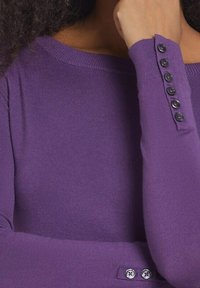 Lila stickad långärmad topp med ett off-shoulder halslinje och fem svarta knappdetaljer på ärmen för både detalj och knäppning.