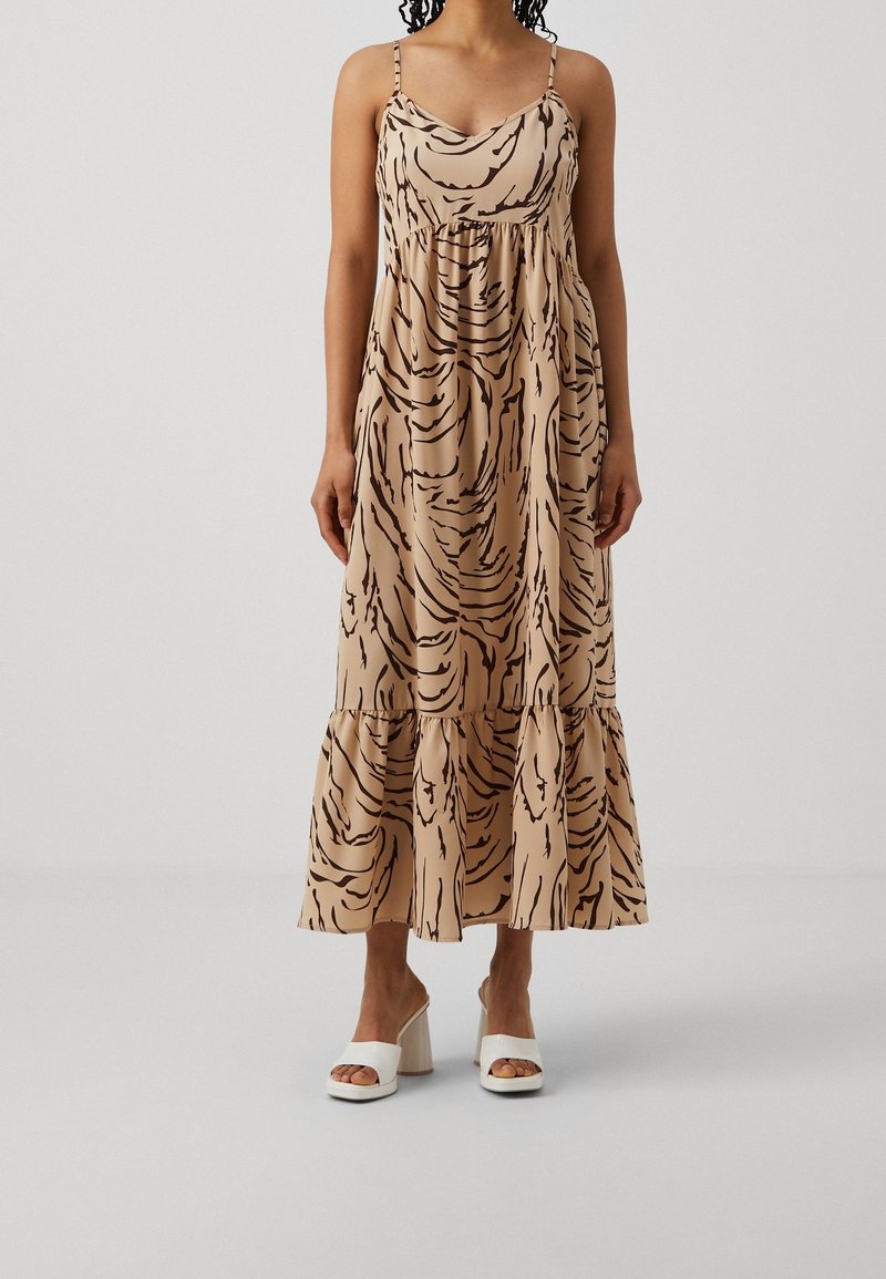 Vestido maxi beige con un patrón negro ondulado, tirantes de espagueti y cintura fruncida. Presenta un dobladillo en capas y se lleva con sandalias blancas de tacón bloque.