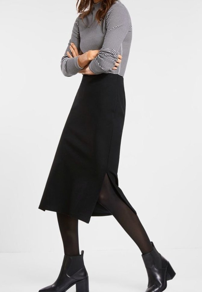 Femme croisant les bras, portant une jupe midi noire fendue sur le côté, un haut à manches longues rayé, des collants noirs et des bottines noires.