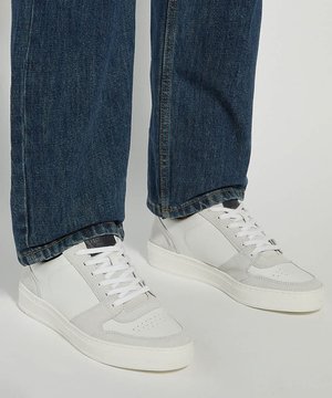 Persoon die witte lage sneakers met veters en een blauwe spijkerbroek draagt, staat op een lichte ondergrond.