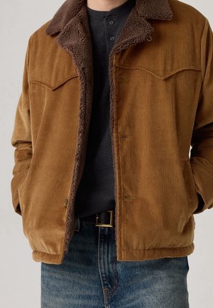 Braune Cordjacke mit dunkelbraunem Fleecefutter, getragen über einem schwarzen Henley-Shirt und blauen Jeans mit schwarzem Gürtel.