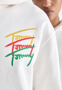 Sudadera blanca con un gráfico texturizado multicolor en el lado izquierdo del pecho, con letras en estilo pincel de color verde, amarillo y rojo.
