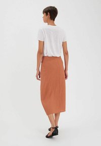 Une jupe en lin de couleur rouille, de longueur mi-longue, au design simple avec une coupe droite et une couture à l'arrière, associée à un t-shirt blanc.