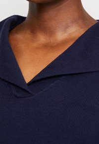 Navy blue knit sweater featuring a deep V-neck and wide collar, with a textured fabric and clean seams. Close-up view of the neckline.