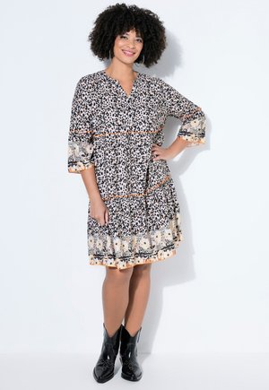 Smilende kvinde med krøllet hår iført en knælængde kjole med dyreprint og blomsterkant og sorte ankelstøvler, stående med hånden på hoften.