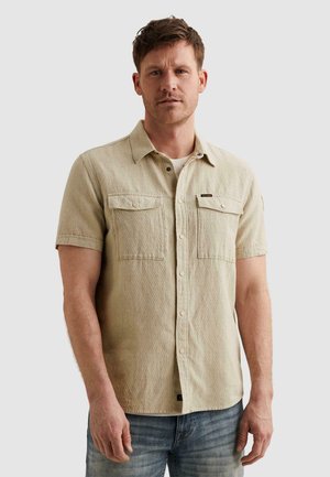 Homme portant une chemise beige texturée à manches courtes avec deux poches poitrine et un jean bleu, debout devant un fond clair uni.