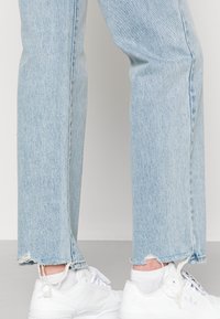 Calças de ganga azul claro com um corte descontraído, bainha desfiada e borda crua. Combinadas com sapatilhas brancas e meias brancas.