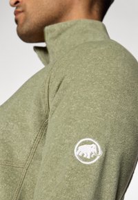 Pullover in pile di un morbido verde oliva, caratterizzato da un colletto con zip e un logo ricamato bianco di un elefante sulla manica sinistra.
