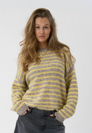 Kvinde med langt bølget hår iført en gul og gråstribet fluffet sweater og mørke bukser, hænderne i lommerne, neutral baggrund.