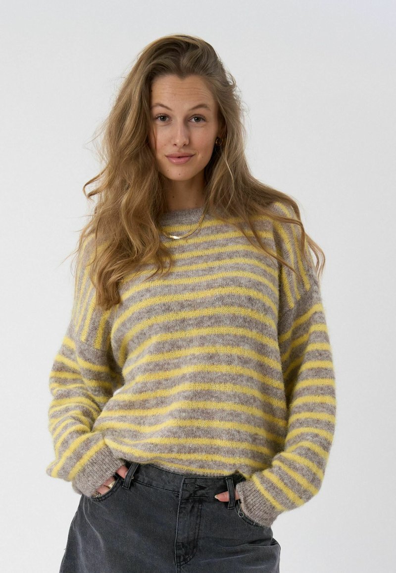 Femme aux cheveux longs et ondulés portant un pull doux à rayures jaunes et grises et un pantalon foncé, les mains dans les poches, fond neutre.