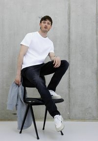 Camiseta blanca de manga corta, jeans negros, sudadera gris colgada sobre una silla negra y zapatillas blancas con detalles verdes contra una pared gris.