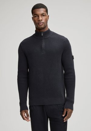 Mann trägt einen dunkelblauen, strukturierten Strickpullover mit Viertel-Reißverschluss-Kragen und schwarze Hose, steht vor einem schlichten hellen Hintergrund.