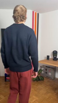 Pull en maille bleu marine avec poignets côtelés, associé à un pantalon rouge à coupe décontractée. En arrière-plan, une table en bois et une plante en pot.