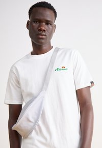 Camiseta blanca de algodón con el logo verde de "ellesse"; bolso cruzado de tela gris con textura suave y correa ajustable que se lleva sobre el hombro.
