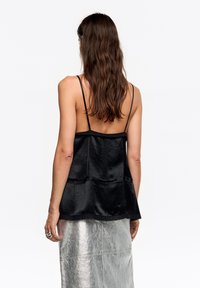 Czarny top bez rękawów z cienkimi ramiączkami i warstwowym fasonem, wykonany z teksturowanej tkaniny z falowanym wykończeniem na dole. Modelka ma na sobie metaliczną spódnicę.