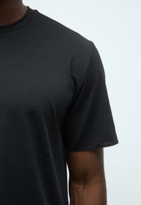 T-shirt noir à manches courtes en tissu lisse. Il présente un col rond et une texture subtile avec un design minimal et sans embellissements visibles.