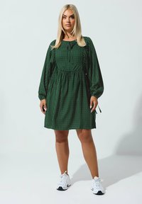 Vihreä ruutukuvioinen mekko, jossa on pitkät hihat, vyötäröä korostava malli ja nauhakiinnitys; malli käyttää valkoisia tennareita ja seisoo vaaleaa taustaa vasten.