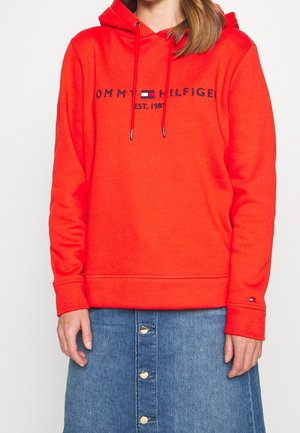 Kobieta w jaskrawoczerwonej bluzie z kapturem Tommy Hilfiger z logo i niebieskiej dżinsowej spódnicy z guzikami, stojąca na tle jednolitego tła.