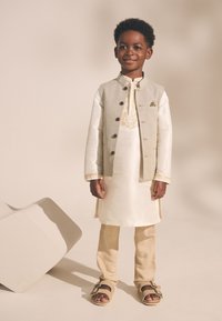 Garçon portant un gilet beige sur un kurta blanc, un pantalon beige clair et des sandales fauves. Détaille une broderie complexe sur le kurta.