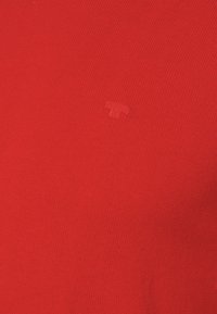 Maglione rosso lavorato a maglia con scollo rotondo e un piccolo logo in rilievo al centro. La texture è liscia con una leggera elasticità.