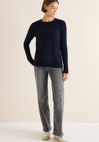 Navyblauer Strickpullover mit langen Ärmeln, rundem Ausschnitt und strukturierten seitlichen Details, kombiniert mit hellgrauen geraden Jeans und Sneakers.