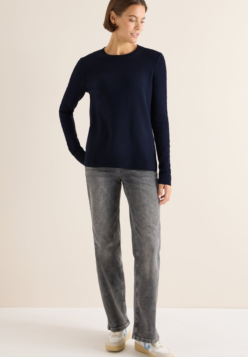 Navyblauer Strickpullover mit langen Ärmeln, rundem Ausschnitt und strukturierten seitlichen Details, kombiniert mit hellgrauen geraden Jeans und Sneakers.