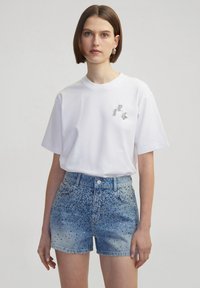 Witte katoenen t-shirt met korte mouwen, versierd met zilveren letters "I", "P" en "K" op de borst, gecombineerd met blauwe denim shorts versierd met studs.