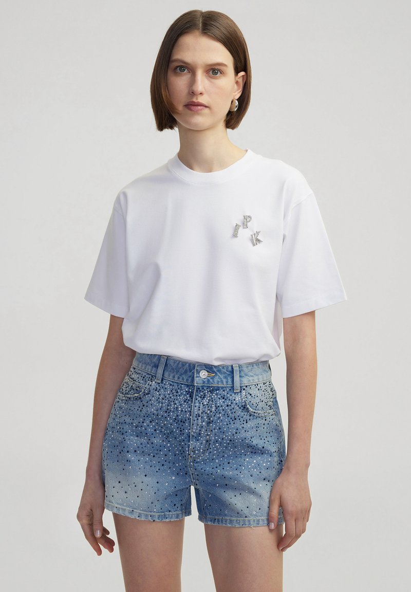 Witte katoenen t-shirt met korte mouwen, versierd met zilveren letters "I", "P" en "K" op de borst, gecombineerd met blauwe denim shorts versierd met studs.