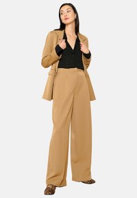Tan blazer op maat over een zwarte blouse met subtiele versieringen, gecombineerd met brede tan broek en schoenen met luipaardprint.