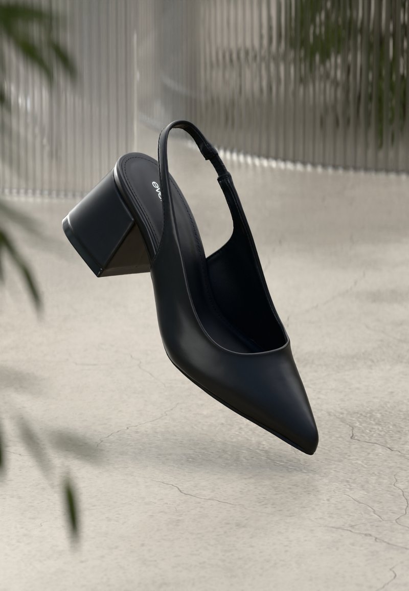 Chaussure slingback en cuir noir avec bout pointu et talon bloc épais sur une surface texturée gris clair.